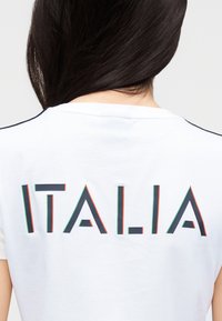 Balta kokvilnas T-krekls ar īsām piedurknēm un uzrakstu "ITALIA" uz muguras biezās, ģeometriskās burtiem ar sarkaniem un zaļiem apmales elementiem.