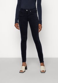 Jean en velours côtelé bleu foncé avec un design ajusté, cinq poches et un accent en bouton métallique au niveau de la taille. Texture lisse, longueur cheville.