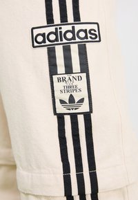 Tkanina boje kreme s crnim adidas logotipom i_oznaku. Sadrži tri vertikalne crne pruge i kontrastni bijeli simbol.