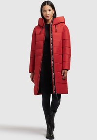 Vrouw in een lange rode gewatteerde jas met een capuchon, zwarte leggings, een zwarte jurk en zwarte enkellaarsjes, die vooruit loopt op een lichte achtergrond.