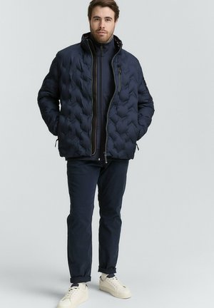 Man draagt een marineblauw doorgestikt jack over een trui met rits, donkere broek en witte sneakers, staat met handen in de zakken van het jack.