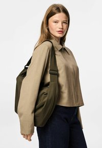 Oliwkowa torebka na ramię wykonana z miękkiej tkaniny, wyposażona w regulowane paski. Modelka ma na sobie beżowy top i ciemne dżinsy, z prostym fasonem kołnierza.