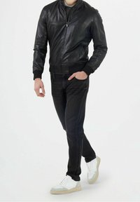 Giacca bomber in pelle nera con polsini a costine, indossata sopra una maglia grigia, abbinata a jeans scuri e sneakers bianche con dettagli grigi.