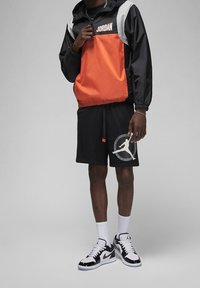 Giacca a vento nera e arancione con logo "JORDAN", abbinata a pantaloni corti neri con una grafica di basket grigia. Calze bianche e scarpe da ginnastica nere.