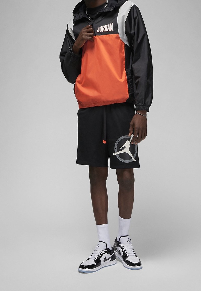 Giacca a vento nera e arancione con logo "JORDAN", abbinata a pantaloni corti neri con una grafica di basket grigia. Calze bianche e scarpe da ginnastica nere.