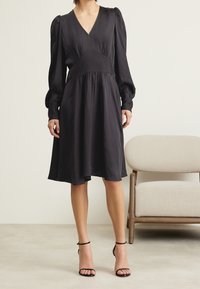 Svart V-ringad klänning med puffärmar, en åtsittande bodice och en luftig kjol. Slätt tyg, knälång, i kombination med svarta remsandaler.