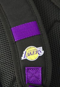 Tecido preto texturizado com uma alça de nylon roxa. Apresenta um logotipo redondo dos Los Angeles Lakers com uma bola de basquete e texto.