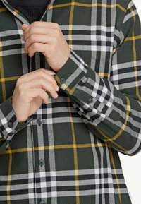 Camicia a quadri verde con controlli neri, bianchi e gialli. Presenta un design con bottoni, maniche lunghe e dettagli a contrasto sui polsini.