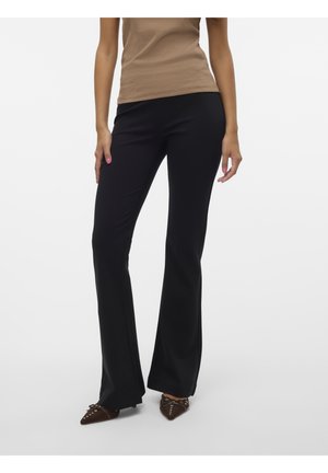 Mujer que lleva pantalones negros ajustados con campana combinados con zapatos marrones puntiagudos y una camiseta sin mangas beige, de pie sobre un fondo blanco.
