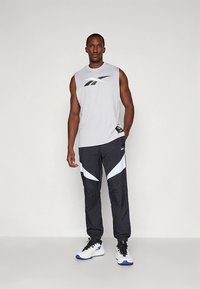 Jasnoszary bezrękawnik z czarnym logo, w połączeniu z czarnymi spodniami dresowymi z białymi akcentami i luźnym krojem. Białe buty sportowe.