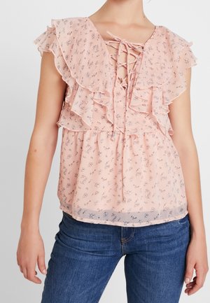 Ærmeløs pink bluse med blomsterprint, flæsekrave og snører foran, kombineret med blå denimjeans på en person.