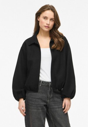 Femme aux longs cheveux bruns portant une veste noire à fermeture éclair, un haut blanc et un jean noir délavé taille haute, debout devant un fond uni.