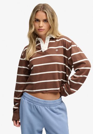 Blonde vrouw met een bruin-wit gestreept cropped shirt met lange mouwen en lichtblauwe joggingbroek, met één hand op haar heup.