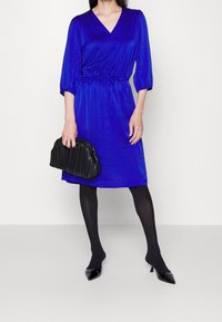 Vestido azul real a la altura de la rodilla con escote en V y cintura ceñida; combinado con un bolso de mano negro plisado y medias negras opacas. Tacones negros puntiagudos.