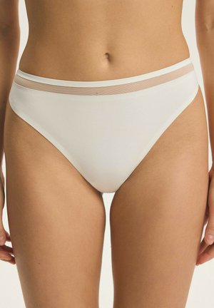 Kobieta ubrana w białą bieliźnę bikini z przeźroczystą siateczkową opaską wokół talii na jednolitym tle.