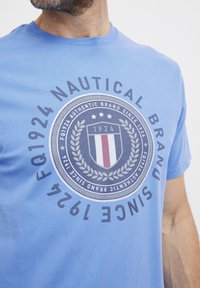 Lyseblå bomullstrøye med et sirkulært grafisk design som har et skjold, stjerner og teksten "NAUTICAL BRAND SINCE 1924" i mørk grå.