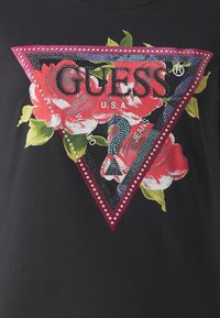Juodas marškinėlis su trikampiu gėlių grafiku, kuriame užrašyta "GUESS" didelėmis raidėmis, papuoštas cirkoniais ir kontrastingomis spalvomis.