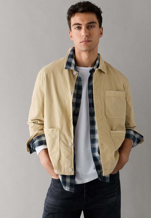Jeune homme aux cheveux foncés portant une veste beige par-dessus une chemise à carreaux et un t-shirt blanc, debout les mains dans les poches devant un fond gris.