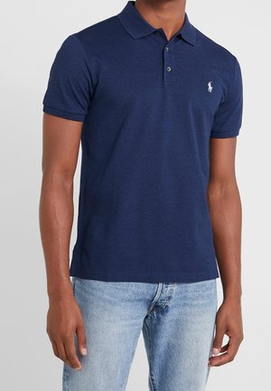 Mand iført navyblå kortærmet polo shirt med lille hvidt logo på brystet og lyseblå cowboybukser, stående mod en ensfarvet baggrund.