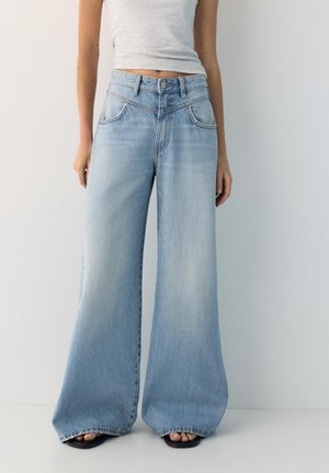 Kvinde iført lyseblå højttaljede vidde jeans og en grå kort top, stående barfodet mod en ensfarvet hvid væg.