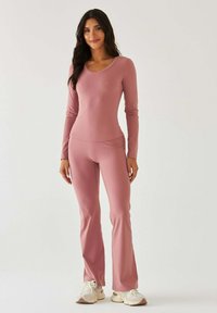 Dopasowany top z długim rękawem i rozkloszowane spodnie z miękkiego, różowego materiału. Gładka faktura z dekoltem w serek. Połączone z jasnymi, kolorowymi sneakersami.