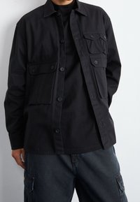 Zwart overhemd met knoopsluiting en een klassieke kraag, twee voorkoteletten en een losse pasvorm, gemaakt van stevig katoen. Gecombineerd met donkere cargobroek.