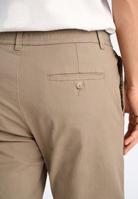 Beige katoenen broek met een slim fit, voorzien van een knoopzak op de achterkant en een gladde textuur. Subtiele horizontale lijnen in de stof.