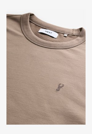 Beige puuvillane T-särk ümarate kaelustega. Sellel on rinnal väike, tikitud homaarimotiiv. Kaeluses on brändi sildike.