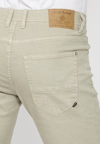 Gros plan sur la poche arrière d'un jean en denim beige avec un patch en cuir de la marque inscrit « Koroshi Brand » et des rivets métalliques aux coins de la poche.
