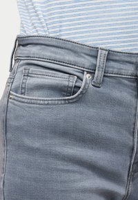 Κοντινό πλάνο τσέπης και θηλιάς ζώνης από ανοιχτό μπλε τζιν τζιν, φορεμένα με λευκό πουκάμισο με μπλε ρίγες, μπουκάτο μέσα στο παντελόνι.