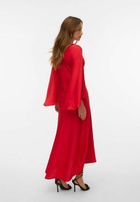 Sarkani, garie piedurkņu maxi kleita, izgatavota no gludas auduma; iezīmē plūstošas zvanu formas piedurknes un brīvu piegriezumu. Valkāta ar melniem papēžu sandalēm.