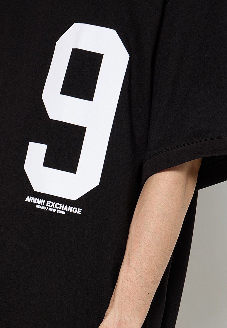 Black oversized t-shirt with a large white number 9 on the back. Features "Armani Exchange" logo in white text at the hem. Cotton material.