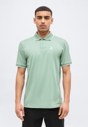 Uomo che indossa una polo a maniche corte verde chiaro con bordi bianchi sul colletto e pantaloni neri, in piedi davanti a uno sfondo semplice.