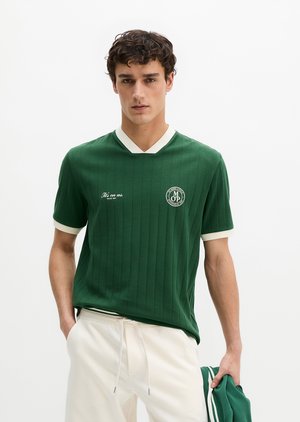 Jeune homme portant une chemise verte à manches courtes avec un bord blanc, un pantalon blanc, tenant une veste verte assortie dans une main.