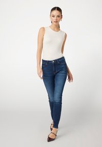 Top sin mangas color beige claro, jeans ajustados azul oscuro y tacones burdeos puntiagudos. Texturas suaves, diseño clásico y silueta ajustada.