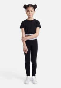 Νεαρή κοπέλα με δύο κότσους, φοράει μαύρο κοντό τοπ και κολάν με λευκές ζώνες Calvin Klein στη μέση και λευκά αθλητικά παπούτσια, στέκεται μπροστά σε λευκό φόντο.