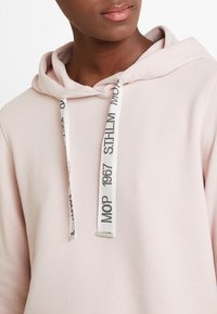 Sudadera con capucha de color rosa claro con cordón y texto blanco contrastante. Textura suave, ajuste relajado, y detalle de bolsillo canguro.