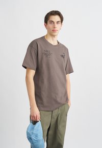 Καφέ βαμβακερό T-shirt με κοντά μανίκια, που διαθέτει μαύρη γραφική εκτύπωση και κείμενο. Το μοντέλο κρατάει ένα ανοιχτό μπλε καπέλο μπέιζμπολ.