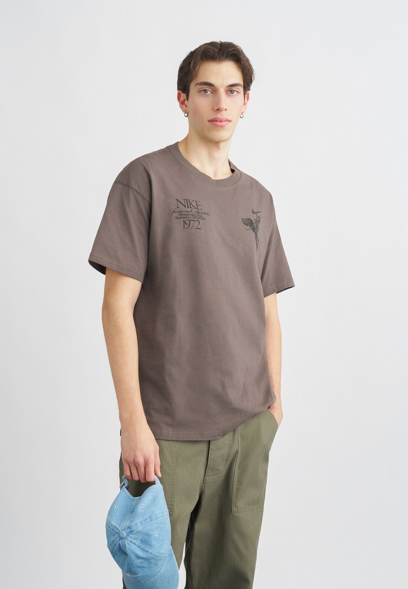 Καφέ βαμβακερό T-shirt με κοντά μανίκια, που διαθέτει μαύρη γραφική εκτύπωση και κείμενο. Το μοντέλο κρατάει ένα ανοιχτό μπλε καπέλο μπέιζμπολ.