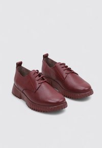 Par de zapatos de cuero de color rojo oscuro con cordones, con parte superior texturada y suela de goma gruesa y acanalada sobre un fondo claro y liso.