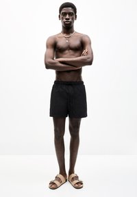 Schwarze Badeshorts mit Kordelzug, kombiniert mit braunen Sandalen mit Schleifchen-Detail. Das Model steht mit verschränkten Armen vor einem weißen Hintergrund.
