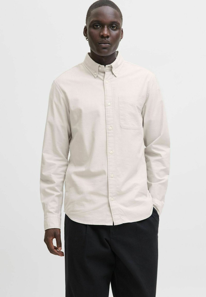 Camicia beige chiaro con bottoni in tessuto di cotone liscio, con taschino sul petto e maniche lunghe, abbinata a pantaloni scuri.