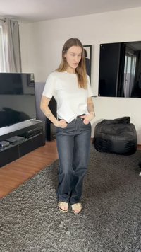 Balta T-krekls, tumši zilas džinsu bikses ar vaļīgu piegriezumu un brūni sandales. Fonā redzams tumšas krāsas TV skapis un sēžammaiss.