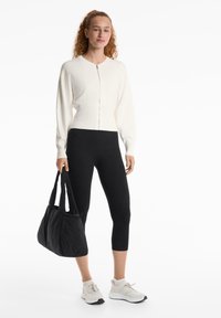 Bijela zip-up cardigan, crne tajice, sive sportske cipele i teksturirana crna torba za rame. Model stoji na jednobojnoj bijeloj pozadini.