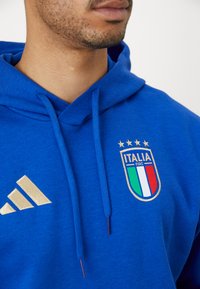Felpa blu realizzata in un materiale morbido, con strisce dorate Adidas e un logo della nazionale italiana ben visibile con tre stelle sopra.