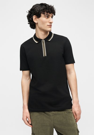 Νέος άνδρας με σγουρά μαλλιά, φοράει μαύρο πόλο με κοντά μανίκια και ριγέ φερμουάρ στον γιακά, καθώς και παντελόνι από βελουτέ ύφασμα σε ελιά πράσινο, κοιτάζει προς τα πλάγια.