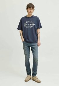 Marineblå bomulls T-skjorte med rund hals og hvit logo print. Båret med lyseblå jeans og beige sko mot en nøytral bakgrunn.