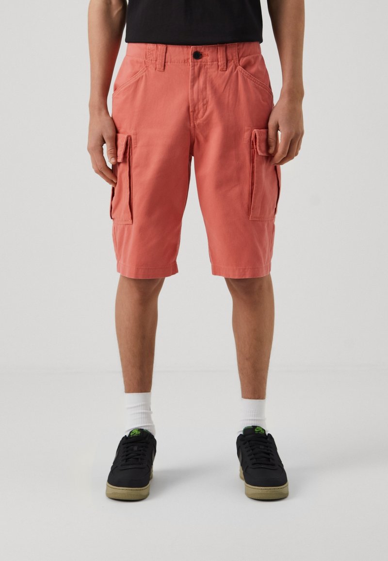 Personne portant un short cargo corail, des chaussettes blanches et des baskets noires, debout devant un fond uni gris clair.