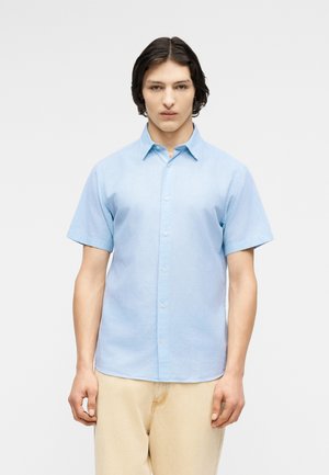 Νέος άνδρας με μεσαίου μήκους σκούρα μαλλιά, φορώντας ανοιχτό μπλε πουκάμισο με κοντά μανίκια και μπεζ παντελόνι, μπροστά σε απλό φόντο.