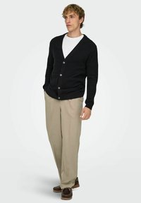 Mustast kudumist kardigan sügava V-kaelusega, millel on viis nuppu, kombineeritud beežide pükste ja pruunide kingadega. Tekstuur tundub pehme ja sile.